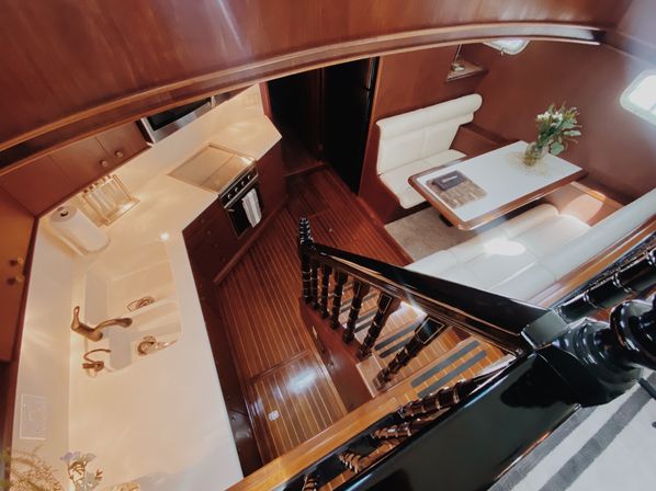 Wood-paneled yacht interior seen from stairs: glossy black railing, compact galley with sink and stove, and white-leather dinette with vase of flowers.