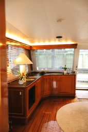 Cozy yacht interior with glossy teak floor, curved cherrywood cabinets, table lamp and potted plants under warm LED ceiling lighting and window blinds