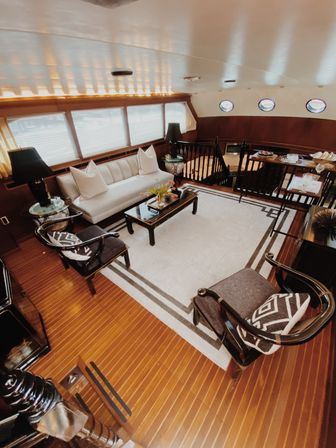 Sunlit luxury yacht salon with white sofa, patterned armchairs, black coffee table on a geometric rug, warm teak plank floors and round porthole windows