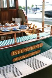 Wooden vintage boat docked at a marina with green-and-white striped bench, outdoor dining table set with navy napkins and glass candleware, waterfront scene in Washington, D.C.