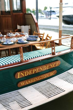 Wooden vintage boat docked at a marina with green-and-white striped bench, outdoor dining table set with navy napkins and glass candleware, waterfront scene in Washington, D.C.