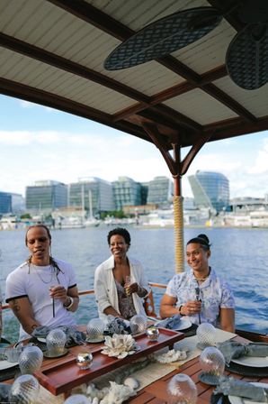 Three friends enjoying drinks around a decorated wooden dining table on a covered boat, with yachts and a modern waterfront skyline in the background