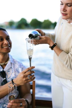 Smiling person holding a champagne flute while another pours bubbly from a bottle on a waterfront boat, sparkling wine and blurred lake treeline in the background