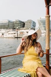 Woman in a white sunhat and yellow sundress sipping wine on a wooden boat at an urban marina with yachts and modern waterfront buildings in the background.