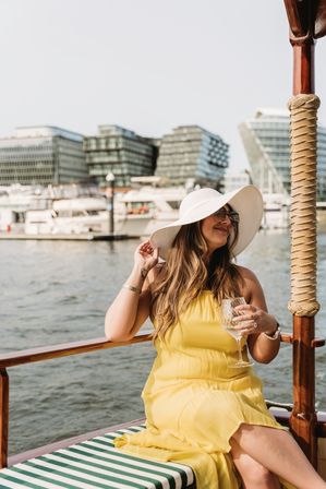 Woman in a white sunhat and yellow sundress sipping wine on a wooden boat at an urban marina with yachts and modern waterfront buildings in the background.