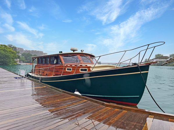 Varnished wooden cabin cruiser with dark green hull moored at a rain‑slick wooden dock on a calm river under a bright blue sky