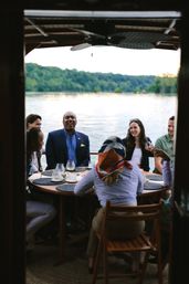 Laughing group of friends enjoying an outdoor lakeside dinner on a boat around a round wooden table with drinks and placemats, wooded shoreline and calm water in the background at dusk