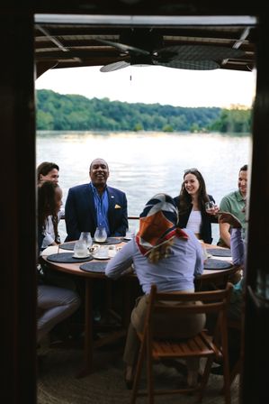 Laughing group of friends enjoying an outdoor lakeside dinner on a boat around a round wooden table with drinks and placemats, wooded shoreline and calm water in the background at dusk