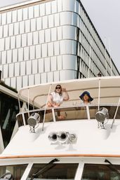 Two women smiling and waving from the upper deck of a luxury yacht beneath a canvas canopy, with a curved modern glass office building rising behind them at an urban waterfront.