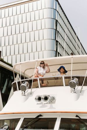 Two women smiling and waving from the upper deck of a luxury yacht beneath a canvas canopy, with a curved modern glass office building rising behind them at an urban waterfront.
