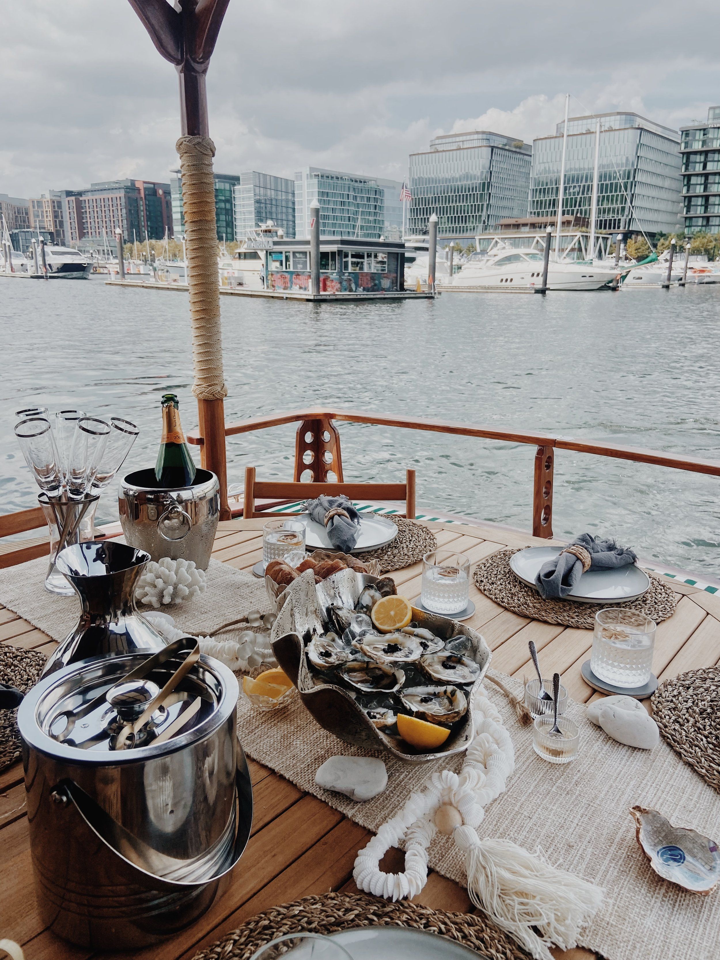 Alfresco nautical table setting: oysters with lemon and chilled champagne on a wooden yacht deck, overlooking an urban marina and glass‑front city skyline.