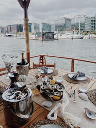 Alfresco nautical table setting: oysters with lemon and chilled champagne on a wooden yacht deck, overlooking an urban marina and glass‑front city skyline.