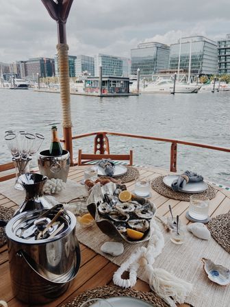 Alfresco nautical table setting: oysters with lemon and chilled champagne on a wooden yacht deck, overlooking an urban marina and glass‑front city skyline.