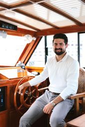 Smiling bearded man in a white shirt seated at the polished wooden helm of a yacht cabin, hand on the ship’s wheel with marina buildings visible through the windows.