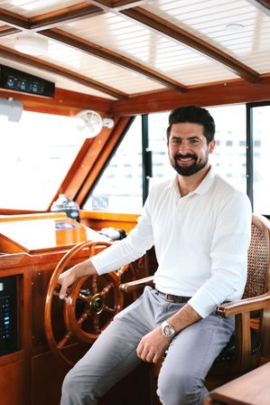 Smiling bearded man in a white shirt seated at the polished wooden helm of a yacht cabin, hand on the ship’s wheel with marina buildings visible through the windows.