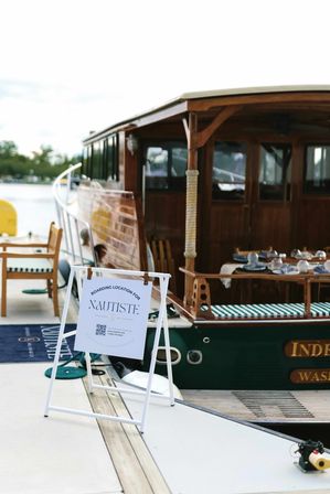 White boarding sign on a marina dock beside a polished wooden boat with striped cushions and a set dining table, ready for a waterfront cruise.