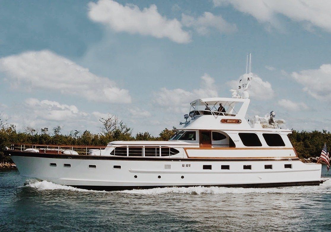 White luxury motor yacht cruising coastal waters under a bright blue sky with fluffy clouds, shoreline trees in the background and an American flag at the stern.