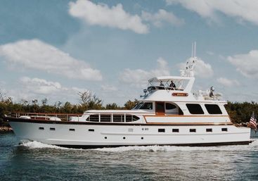White luxury motor yacht cruising coastal waters under a bright blue sky with fluffy clouds, shoreline trees in the background and an American flag at the stern.