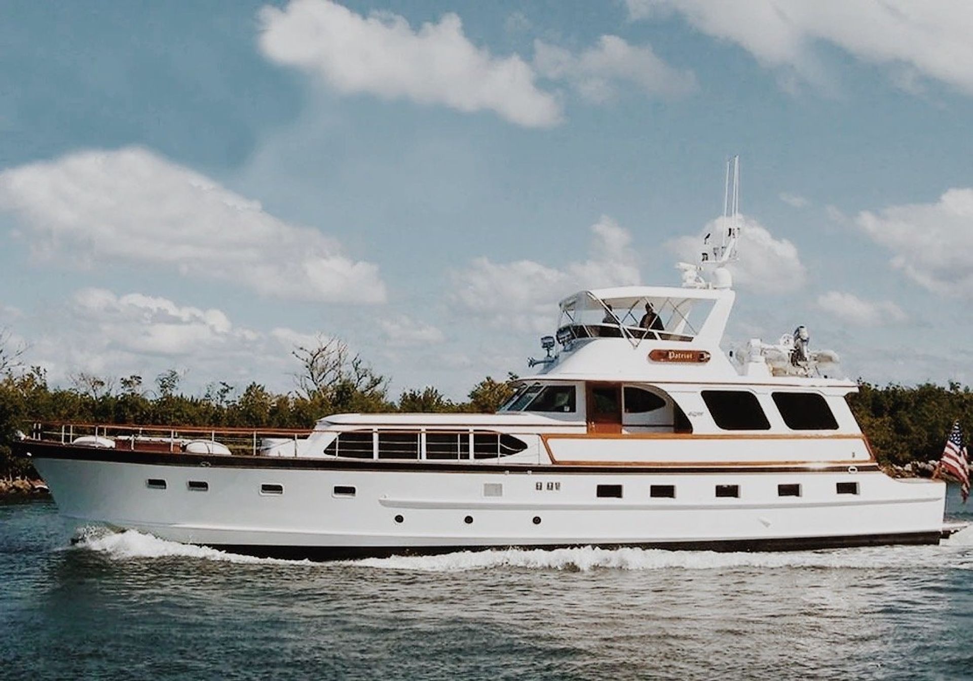 White luxury motor yacht cruising coastal waters under a bright blue sky with fluffy clouds, shoreline trees in the background and an American flag at the stern.