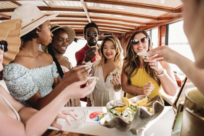 Group of friends on a sunny boat cruise enjoying fresh oysters with lemon over a seafood platter inside a wooden cabin, summer brunch by the ocean