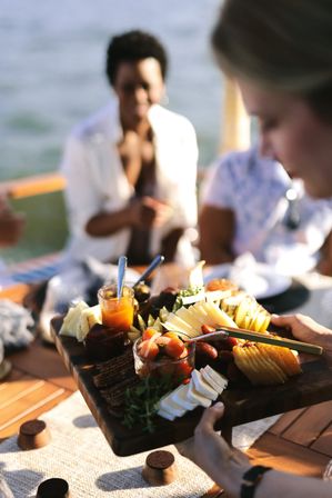 Sunlit wooden charcuterie board with sliced cheeses, crackers, olives, dates, cherry tomatoes and jars of jam being passed among friends on a boat over water