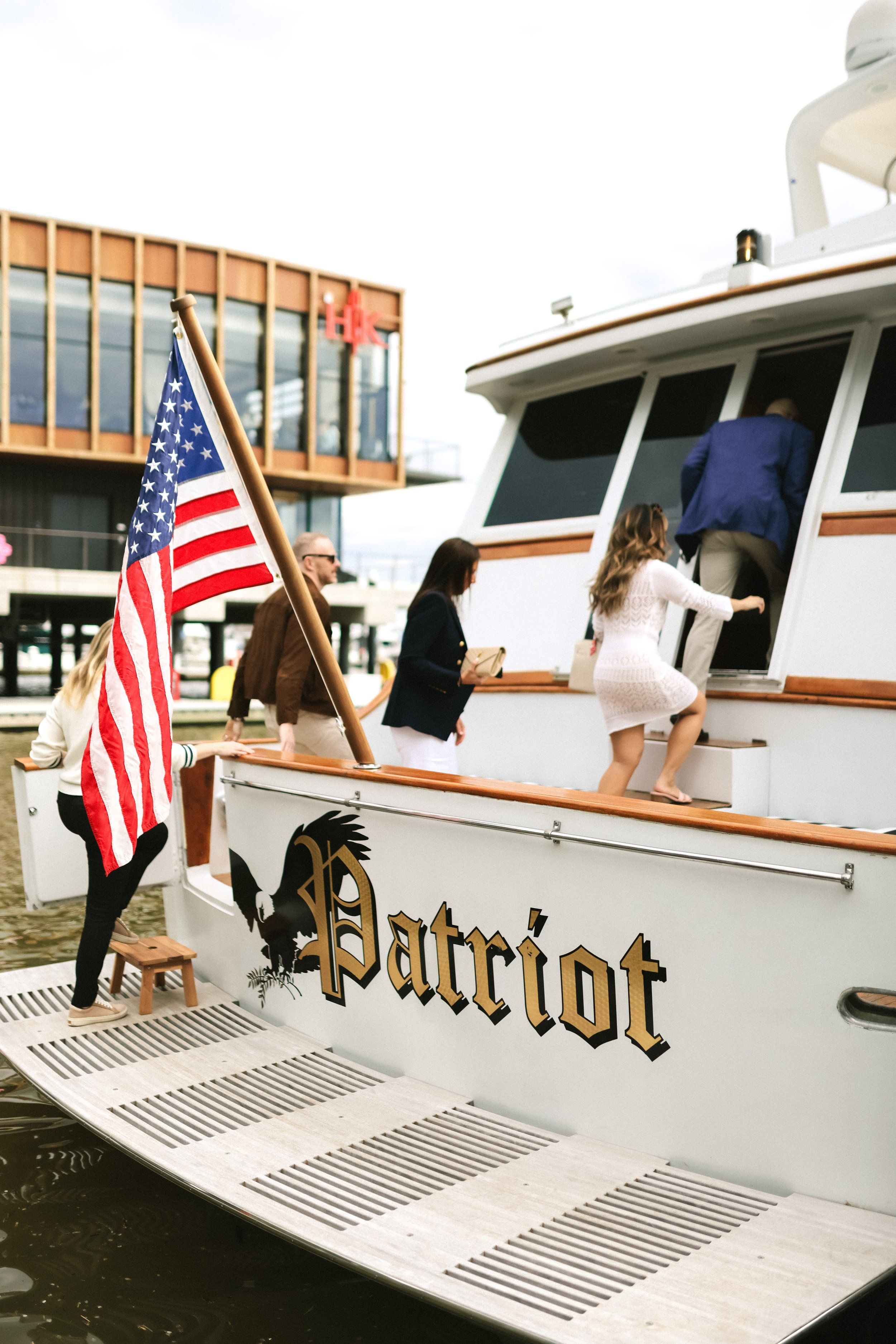 Group boarding a white yacht at a marina, American flag flying from the stern and a modern waterfront building in the background.