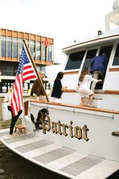 Group boarding a white yacht at a marina, American flag flying from the stern and a modern waterfront building in the background.