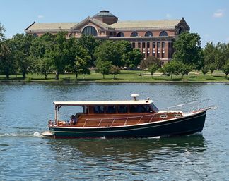 Wooden cabin cruiser gliding on a calm river in summer, passing a tree-lined waterfront park with a large historic red-brick neoclassical building under a clear blue sky.