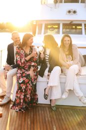Four friends laughing on a yacht deck at sunset, clinking wine glasses in warm golden-hour light — floral dress and casual chic outfits.