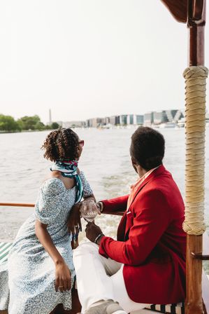 Stylish couple on a sunny river cruise, woman in a floral dress and scarf and man in a red blazer holding a wine glass, gazing at a modern city skyline from a wooden boat
