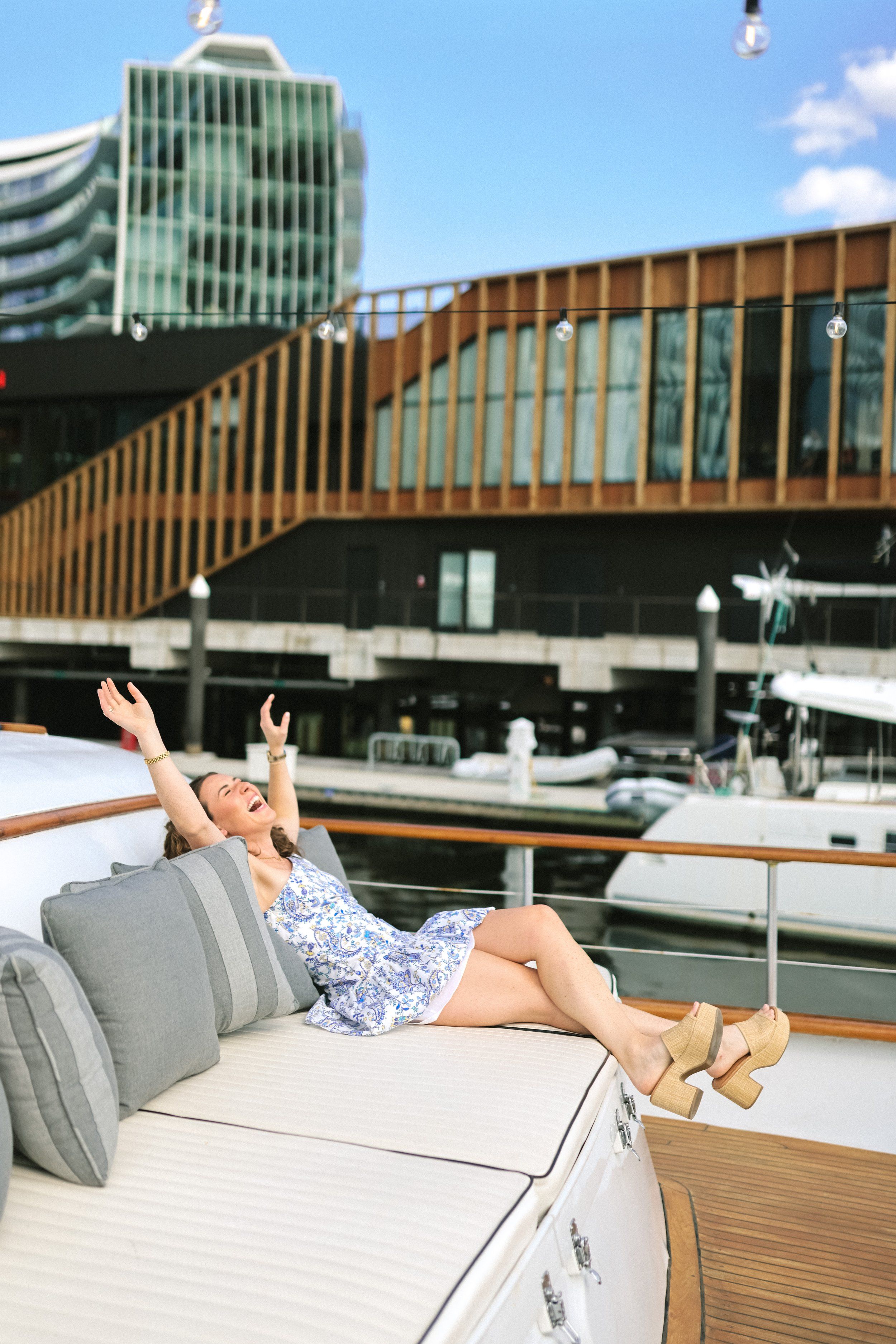 Young woman in a floral sundress lounging on a cushioned yacht deck at a sunny marina, arms raised in joy with modern waterfront buildings and moored boats in the background.