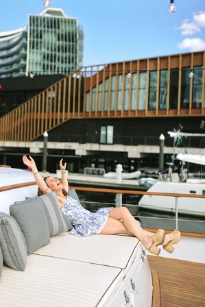 Young woman in a floral sundress lounging on a cushioned yacht deck at a sunny marina, arms raised in joy with modern waterfront buildings and moored boats in the background.