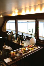 Charcuterie grazing board with cheeses, crackers, fruit and dips on a wooden table inside a cozy boat cabin by bright waterfront windows with wine bottles nearby.