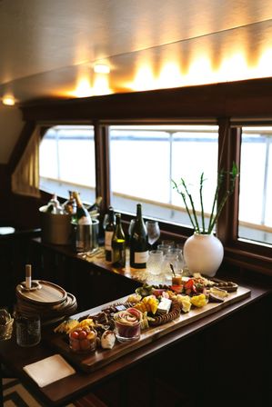 Charcuterie grazing board with cheeses, crackers, fruit and dips on a wooden table inside a cozy boat cabin by bright waterfront windows with wine bottles nearby.