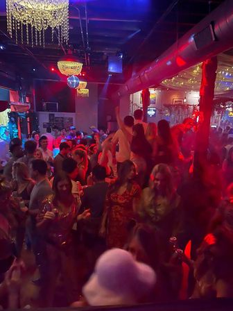 Crowded indoor nightclub dance floor with chandeliers and a disco ball, red and blue lighting and string lights, people dancing and holding drinks in a lively urban nightlife scene.