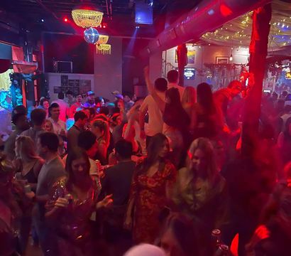 Crowded indoor nightclub dance floor with chandeliers and a disco ball, red and blue lighting and string lights, people dancing and holding drinks in a lively urban nightlife scene.