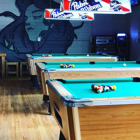 Three green-felt pool tables lined up on a wooden floor in a lively pool hall, racked billiard balls, Pabst Blue Ribbon light fixtures and a mural on the cinderblock wall, ready for a game.
