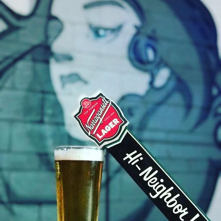 Tall glass of golden lager beneath a red lager tap badge and a Hi-Neighbor tap handle, set against a blurred teal mural of a woman’s face