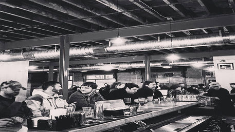 Black-and-white image of a busy urban industrial-style taproom with patrons seated along a long wooden bar, glasses and shakers on the counter, and exposed ceiling beams and metal ductwork overhead.
