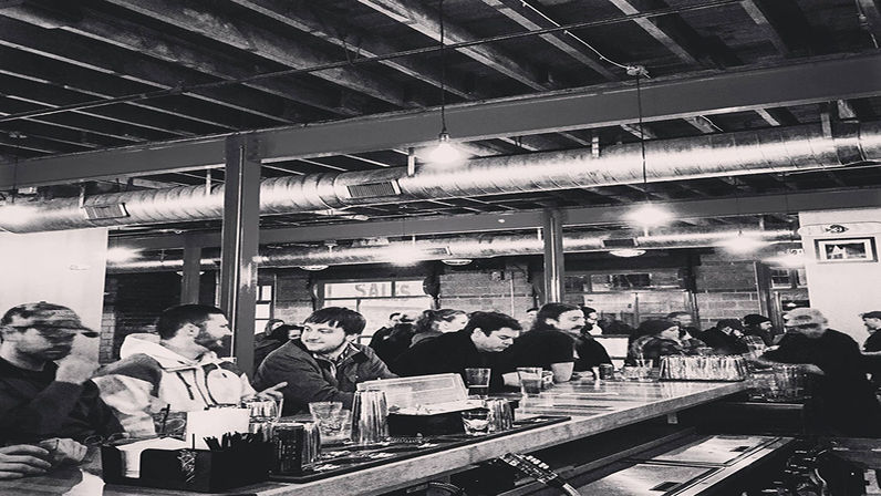 Black-and-white image of a busy urban industrial-style taproom with patrons seated along a long wooden bar, glasses and shakers on the counter, and exposed ceiling beams and metal ductwork overhead.