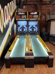 Two adjacent skee-ball arcade lanes with yellow trim and LED-lit ramps, digital scoreboards, exposed brick wall and hardwood floor