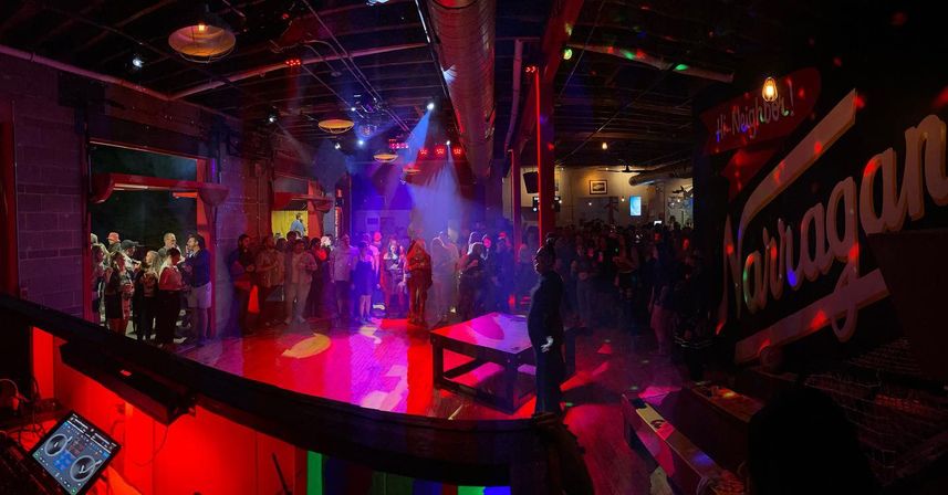 Crowded industrial-style music venue at night with a central dance floor, colorful stage and disco lights, a DJ booth in the foreground, exposed ductwork and a lively standing crowd.