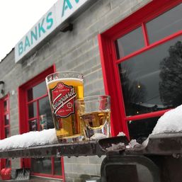 Amber pint and shot glass on a snowy metal railing outside a red‑trimmed pub or brewery, a winter toast against a gray brick facade