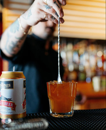 Tattooed bartender stirring an amber craft cocktail over ice with a long bar spoon, a can of ginger beer beside the glass and blurred bar bottles in the background.