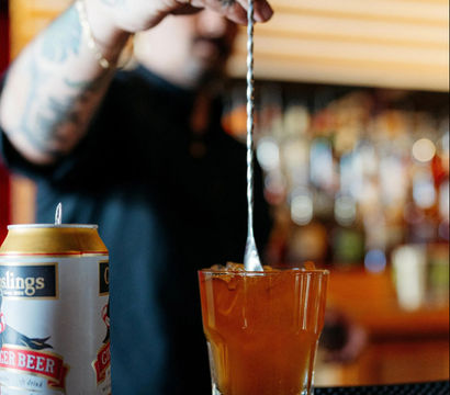 Tattooed bartender stirring an amber craft cocktail over ice with a long bar spoon, a can of ginger beer beside the glass and blurred bar bottles in the background.