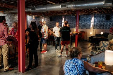 Casual crowd mingling in an industrial-chic indoor lounge with exposed ductwork, red support columns, geometric wallpaper, concrete floor, high-top tables and cozy seating, people chatting and holding drinks.