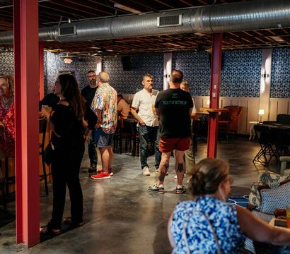 Casual crowd mingling in an industrial-chic indoor lounge with exposed ductwork, red support columns, geometric wallpaper, concrete floor, high-top tables and cozy seating, people chatting and holding drinks.