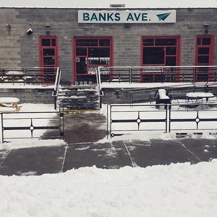 Snowy urban storefront with red-trim windows, empty outdoor patio tables, metal railings and a shoveled sidewalk leading up steps to the entrance beneath a rectangular sign.
