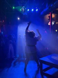 Silhouette of a dancer striking a high-energy pose on a smoke-filled, blue-lit nightclub dance floor under a disco ball, surrounded by a watching crowd and colorful stage lights.