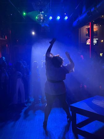 Silhouette of a dancer striking a high-energy pose on a smoke-filled, blue-lit nightclub dance floor under a disco ball, surrounded by a watching crowd and colorful stage lights.